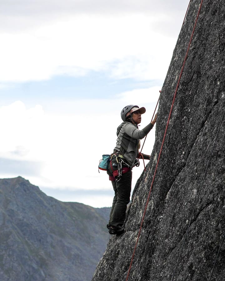 A climber on a granite arete with a sharp green mountain ridge filling the sky behind her