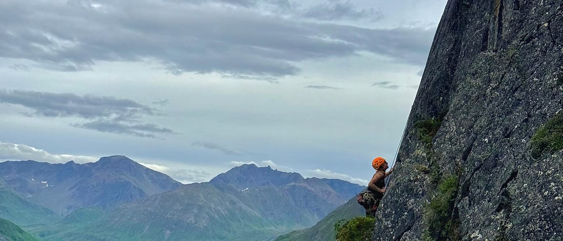 A WCA climber high on a dark buttress above a green Alaskan valley