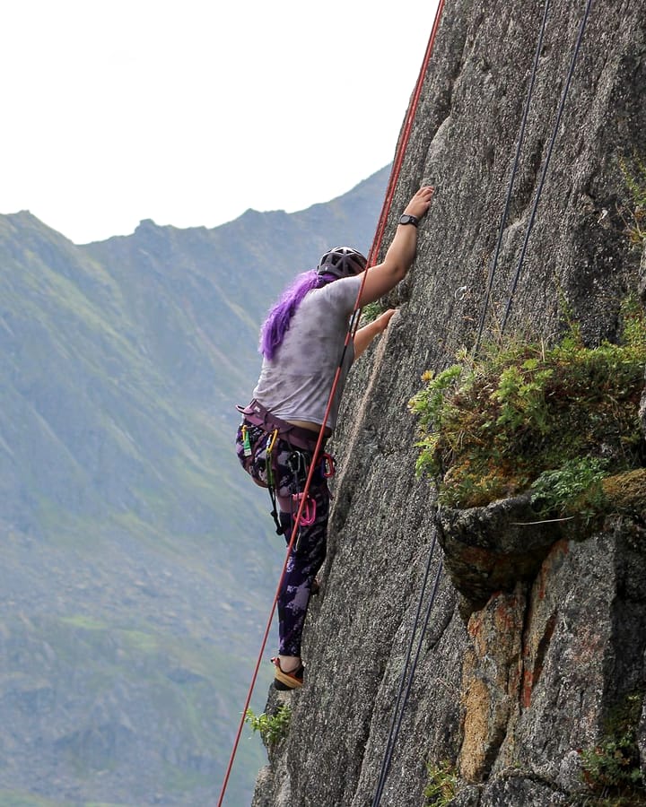 A climber in a magenta top reaches for a hold on grey granite, a long green Alaskan ridge behind her