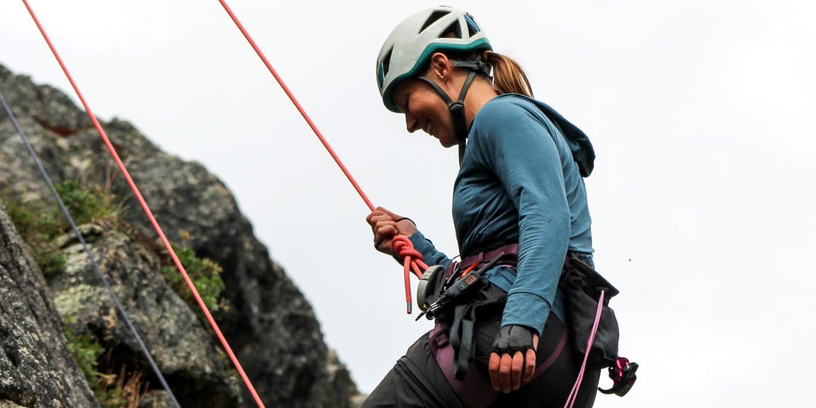 A climber leans back on her rope on a rock ledge and laughs, ropes running past her shoulder