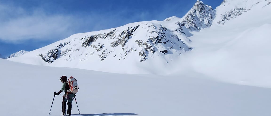 A WCA skier breaks trail alone across a wide snowfield beneath a high Alaskan peak