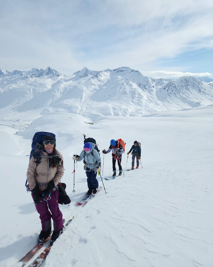 Four skiers travel single file across a snow basin below a long range of white Alaskan peaks
